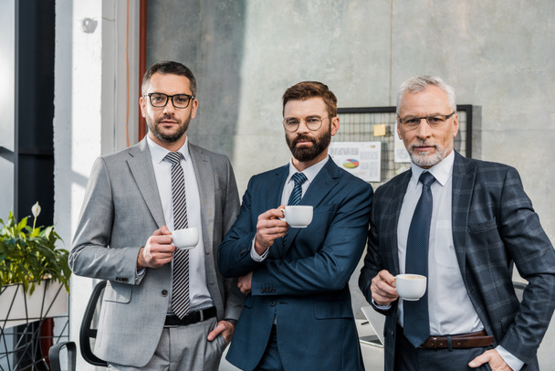 three confident businessmen holding cups of coffee and looking at camera in office - Foto, afbeelding