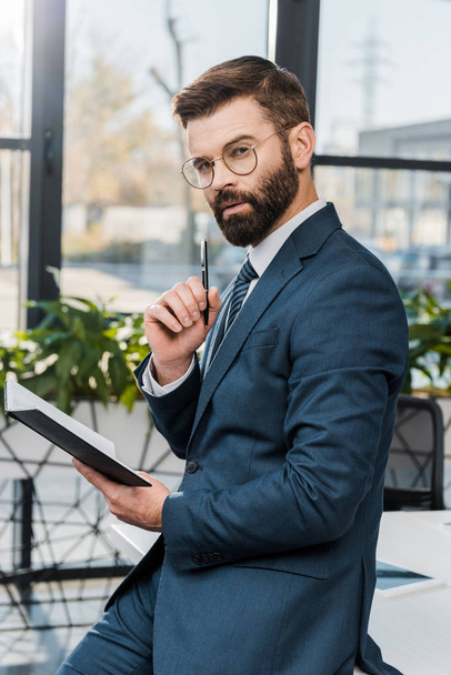 serious bearded businessman holding notepad and looking at camera in office - Φωτογραφία, εικόνα
