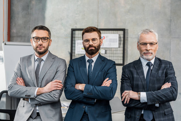 confident businessmen standing with crossed arms and looking at camera in office - Foto, imagen