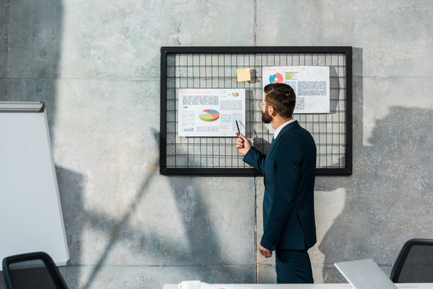side view of bearded businessman holding pen and pointing at business charts on wall - Photo, Image