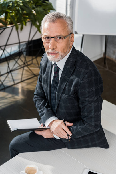 high angle view of serious mature businessman sitting on office table and looking at camera - Zdjęcie, obraz