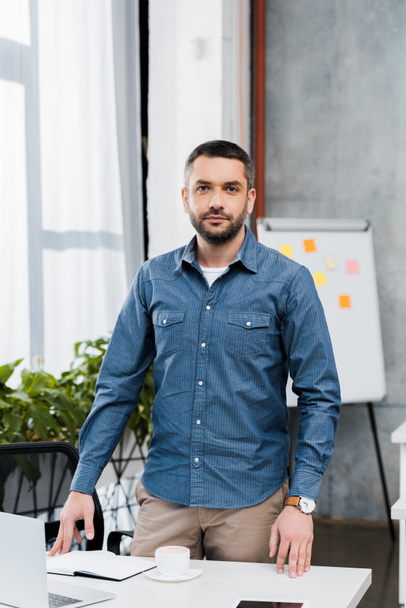 handsome bearded businessman standing at workplace and looking at camera - Fotografie, Obrázek