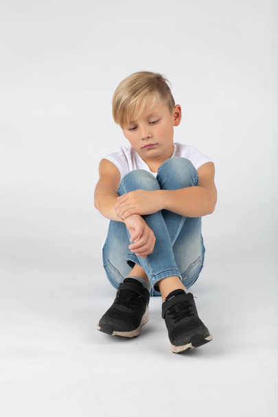 little blond boy is sitting sadly on the floor white background - Foto, immagini