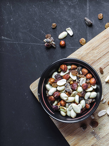 Top view of black bowl with nuts and dried fruits on the wooden cutting board with black chalkboard background. Several types of nuts including hazel-nuts and peanuts. - Foto, imagen
