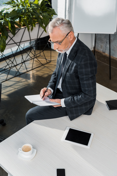 високий кут зору зрілого бізнесмена, що пише в блокноті, сидячи на офісному столі
 - Фото, зображення