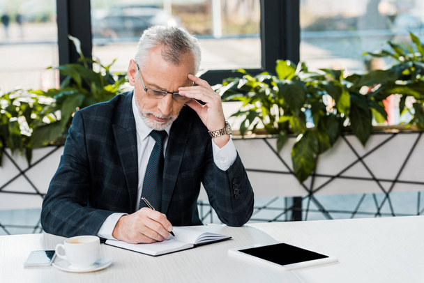 focused mature businessman sitting at workplace and writing in notebook - Foto, afbeelding