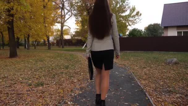 zakenvrouw in rok en jasje met zwarte werkmap in de hand, gaat langs de stoep huis door herfst park - Video
