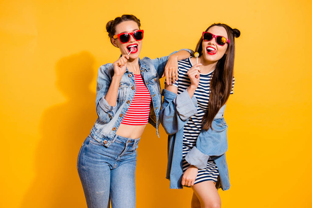 Two brunette hair beautiful attractive dreamy charming lady glasses spectacles casual denim outfit isolated on yellow shine background eating sugar sweet yummy calories caramel confectionery dessert - Фото, изображение