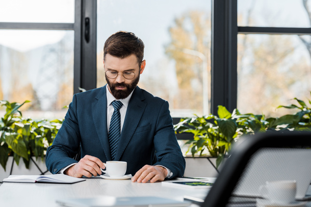 serious bearded businessman in suit and eyeglasses holding cup of coffee while sitting at workplace - Photo, Image
