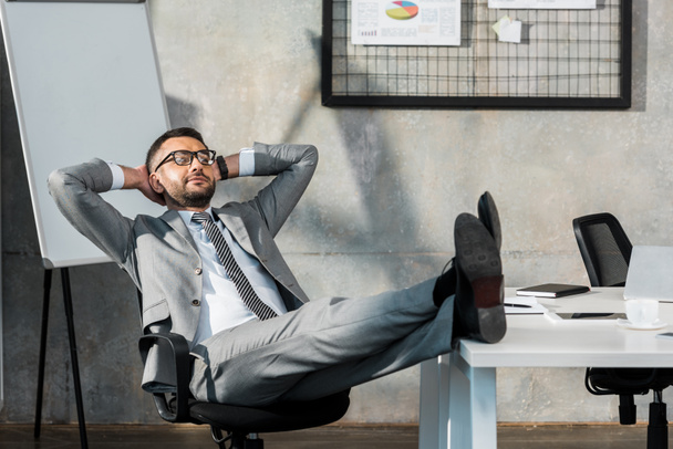relaxed buisnessman in eyeglasses sitting with legs on table and hands behind head in office - Photo, Image