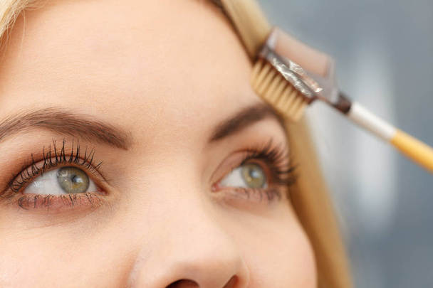 Close up of woman doing her make up, preparing brows using brush tool brushing eyebrows. - Фото, изображение