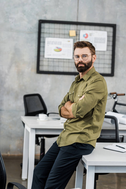 handsome bearded businessman in eyeglasses sitting with crossed arms and looking at camera in office - Photo, Image