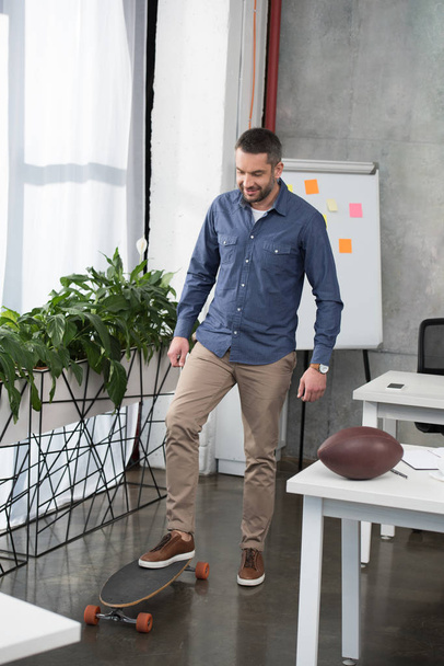 cheerful handsome businessman putting leg on longboard in office - Photo, Image