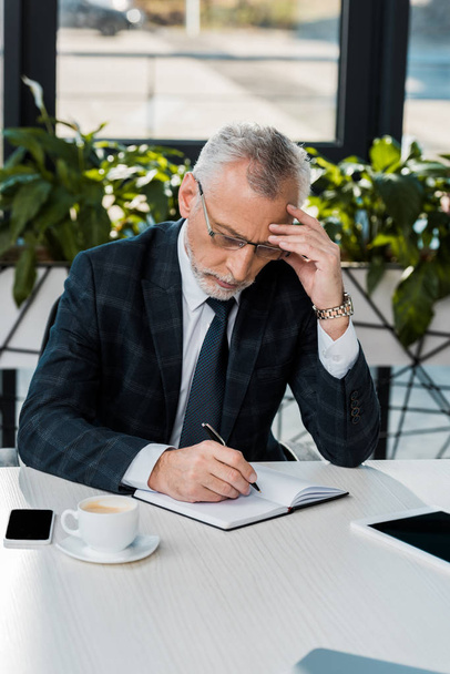 pensive handsome middle aged businessman in suit writing something to notebook in office - Photo, Image