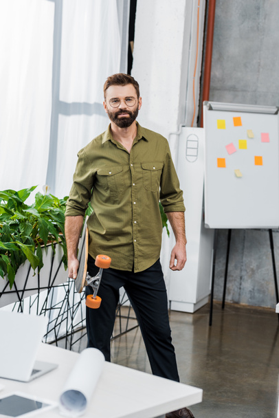 handsome bearded businessman standing with longboard and looking at camera in office - Photo, Image