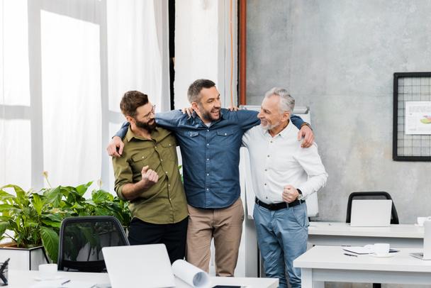 drie glimlachend knappe zakenlieden knuffelen en permanent in kantoor - Foto, afbeelding