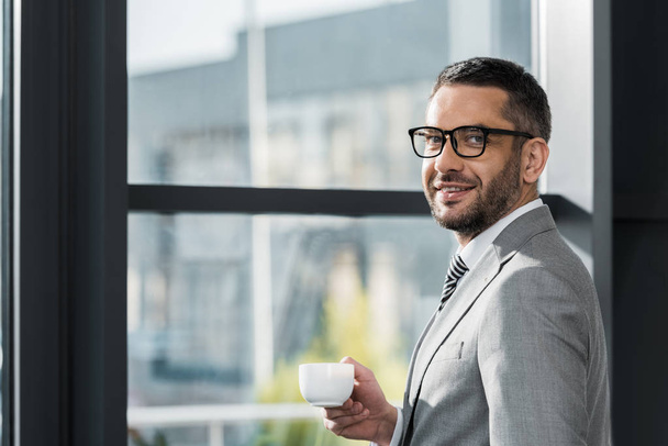 handsome businessman in suit and glasses standing near window with cup of coffee and looking at camera in office - Foto, Imagen