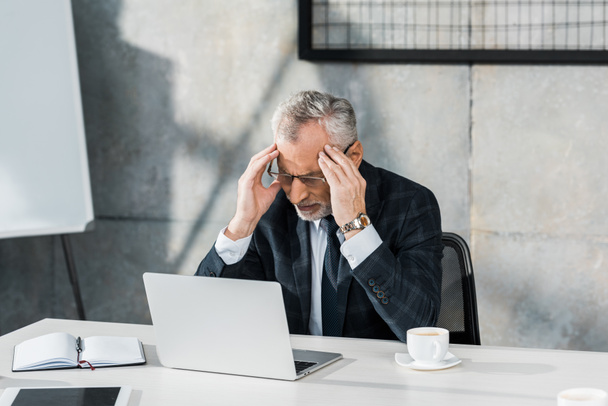 exhausted middle aged businessman touching head at table with laptop in office - Fotoğraf, Görsel