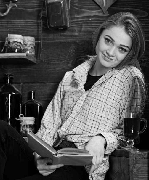 Relaxation concept. Girl in casual outfit sits with book in wooden vintage interior. Lady on smiling face in plaid clothes looks casual. Girl student relaxing with book and glass of mulled wine - Фото, изображение