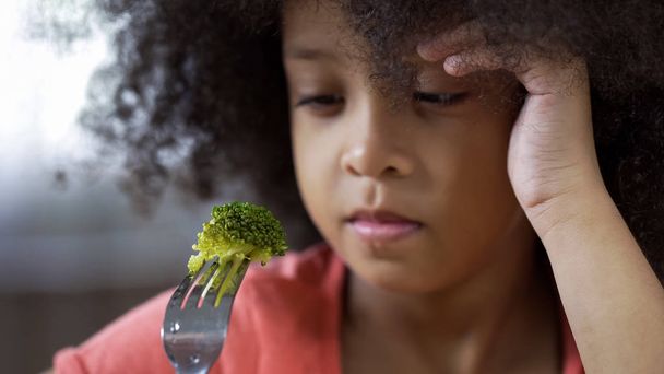 Preschool African girl sadly looking at piece of broccoli on, healthy nutrition - Foto, afbeelding