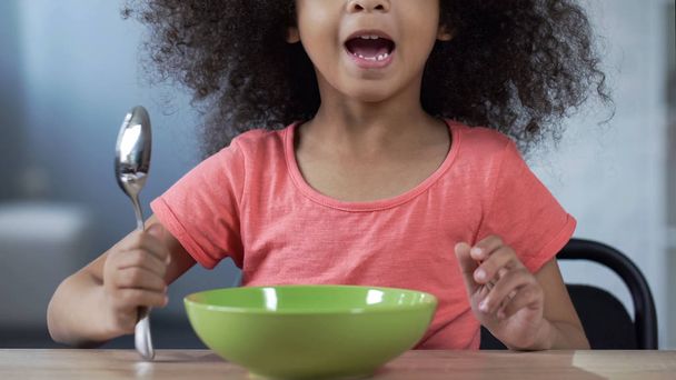 Cute mulatto girl sitting at table with spoon and asking for dinner, hungry kid - Foto, imagen