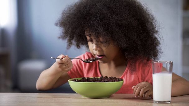 Adorable African girl eating chocolate cornflakes with appetite, drinking milk - Zdjęcie, obraz