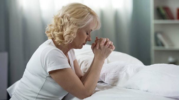 Ανώτερος γυναίκα κάθεται στο πάτωμα κοντά στο κρεβάτι και προσεύχεται, θρησκείας και πεποιθήσεων - Φωτογραφία, εικόνα