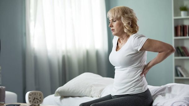 Blond mature lady sitting on bed and touching her back, radiculitis and pain - Φωτογραφία, εικόνα