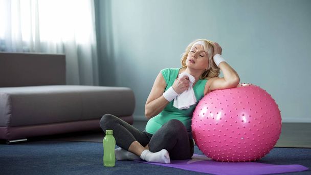 Exhausted active lady wiping with towel after home workout with fitness ball - Фото, изображение