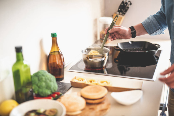 Κοντινό πλάνο της αρσενική χέρι κρατά κουτάλι κουζίνας με ταλιατέλες. Στέκεται κοντά σόμπα - Φωτογραφία, εικόνα