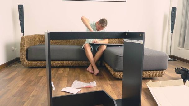 A young man independently collects furniture in the living room of his house. A man collects a computer desk. - Φωτογραφία, εικόνα