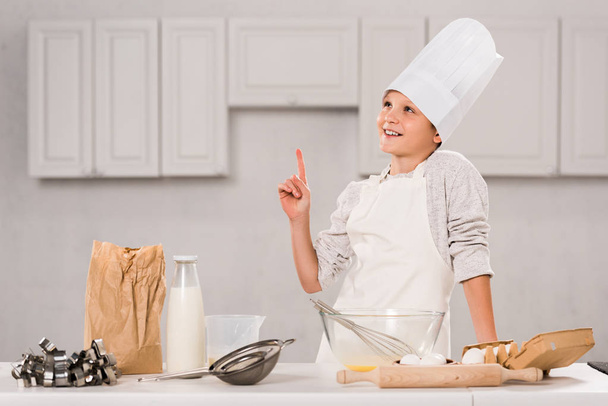 cheerful boy in chef hat and apron doing idea gesture during food preparation at table in kitchen - Fotó, kép