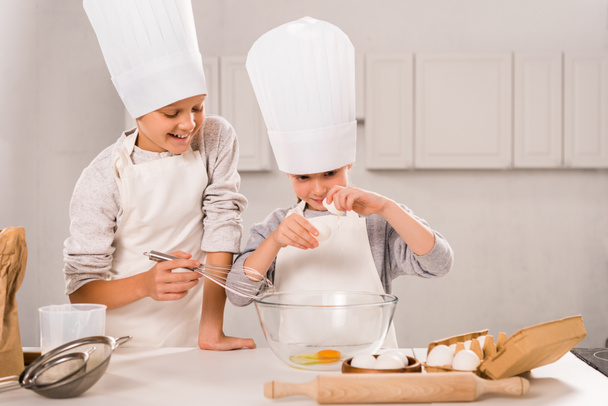 uśmiechający się brat i siostra w fartuchy i czapki szefa kuchni podczas przygotowywania posiłków przy stole w kuchni  - Zdjęcie, obraz