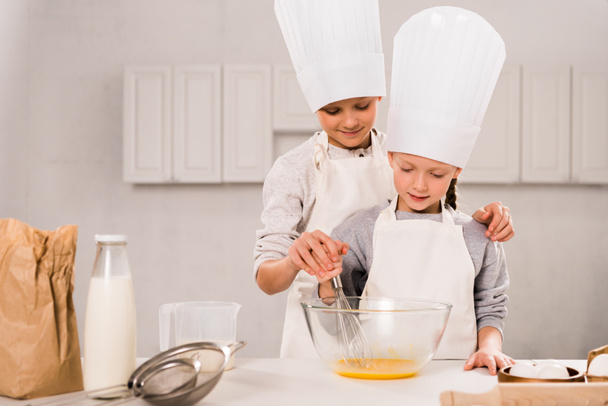 niños en delantales y sombreros de chef batiendo huevos en tazón en la mesa en la cocina
  - Foto, Imagen