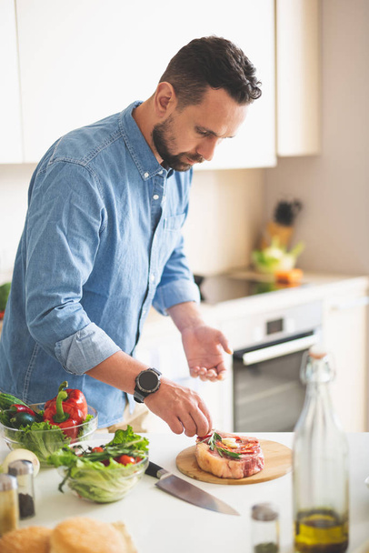 Young man adding herbs to meat while standing near kitchen table - Foto, Imagen