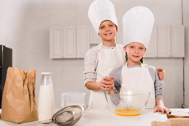 happy sister and brother in aprons and chef hats whisking eggs in bowl at table in kitchen  - Photo, image