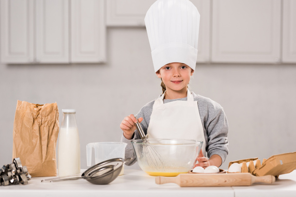 glimlachend kind in chef-kok hoed eieren te zwaaien in kom aan tafel in de keuken - Foto, afbeelding