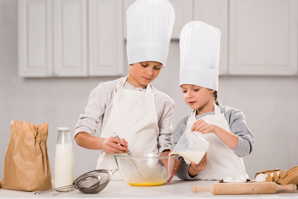 kid pouring milk into bowl while brother whisking at table in kitchen  - Photo, Image