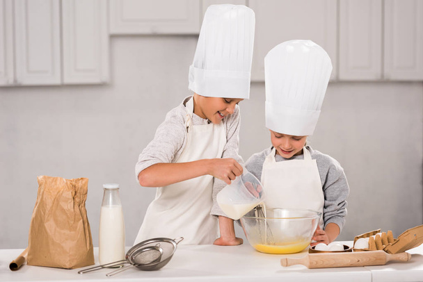 boy pouring milk into bowl while sister standing near at table in kitchen  - Foto, imagen