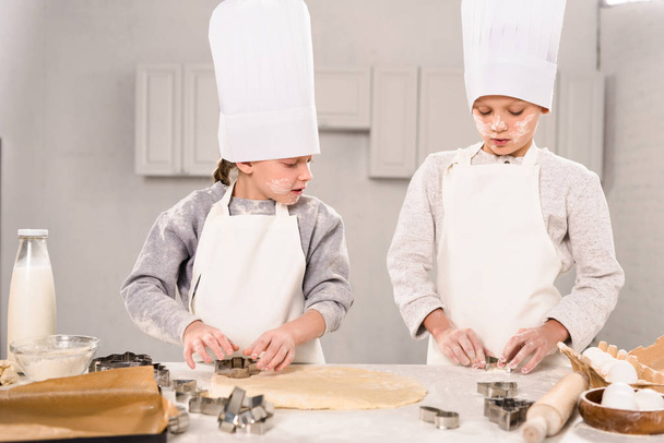 selective focus of little brother and sister in chef hats and aprons cutting out dough for cookies at table in kitchen - Фото, изображение