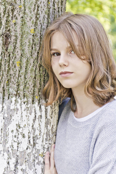 Beautiful girl fourteen years old lean against tree - Фото, изображение