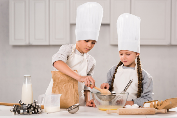 brother and sister in chef hats and aprons whisking eggs in bowl at table in kitchen - Valokuva, kuva