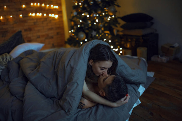 Молоді коханці, покриті ковдрою, лежать в ліжку під час кохання на фоні ялинки та прикрас
. - Фото, зображення