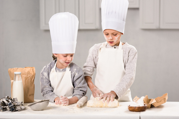 niños en delantales y sombreros de chef haciendo masa con rodillo en la mesa en la cocina
 - Foto, Imagen