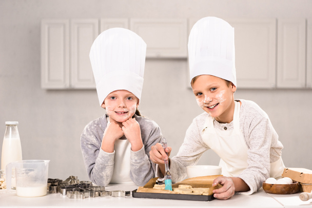 iloiset lapset esiliinoissa harjaamalla evästeitä keittiössä
  - Valokuva, kuva