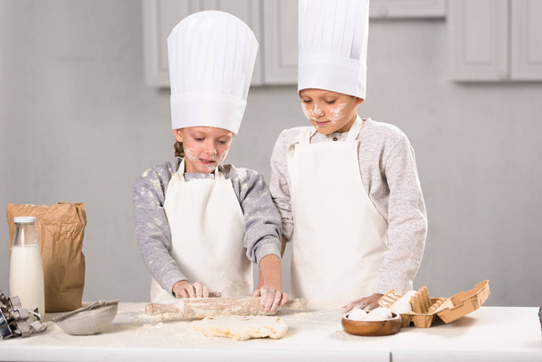 sister and brother in aprons and chef hats making dough with rolling pin at table in kitchen - Foto, Imagem