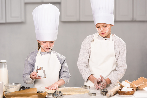 kinderen in chef mutsen en schorten uitsnijden van deeg voor cookies aan tafel in de keuken - Foto, afbeelding