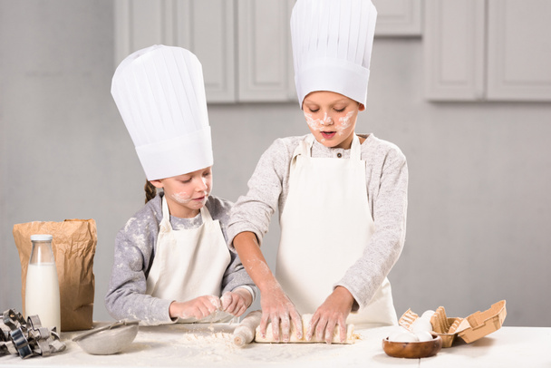 niños enfocados en delantales y sombreros de chef haciendo masa con rodillo en la mesa en la cocina
 - Foto, Imagen