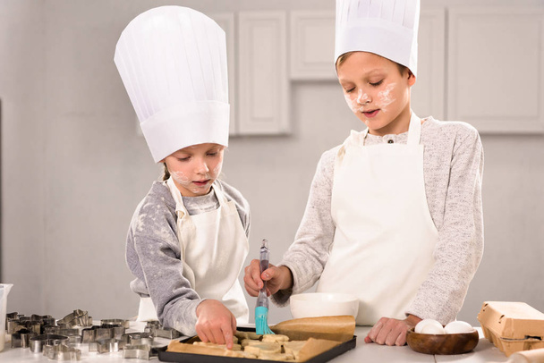 kinderen geconcentreerd in schorten borstelen van cookies op de bakplaat in keuken  - Foto, afbeelding