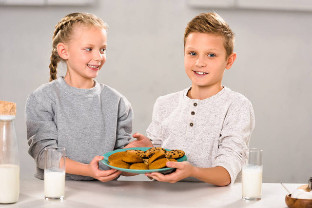 счастливые дети держат тарелку с печеньем возле стола с молочными стаканами
  - Фото, изображение